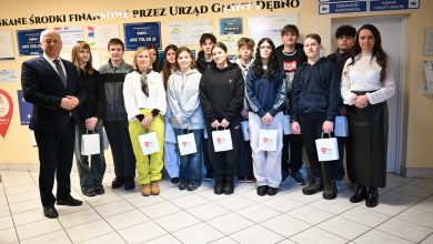 Na zdjęciu widać grupę młodzieży – uczniów wraz z opiekunami – pozujących wewnątrz budynku Urzędu Gminy Dębno. Osoby stoją w jednym rzędzie, część z nich trzyma papierowe torby z logo gminy. Po bokach grupy znajdują się dorośli – kobieta i mężczyzna w formalnych strojach. W tle widoczne są tablice informacyjne oraz plansze z informacjami o środkach finansowych pozyskanych przez urząd. Scena ma oficjalny, ale przyjazny charakter i dokumentuje wizytę młodzieży w urzędzie.