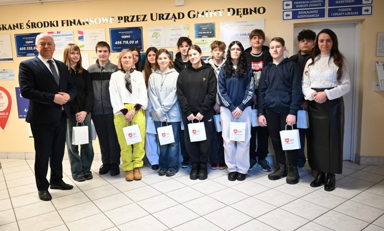 Na zdjęciu widać grupę młodzieży – uczniów wraz z opiekunami – pozujących wewnątrz budynku Urzędu Gminy Dębno. Osoby stoją w jednym rzędzie, część z nich trzyma papierowe torby z logo gminy. Po bokach grupy znajdują się dorośli – kobieta i mężczyzna w formalnych strojach. W tle widoczne są tablice informacyjne oraz plansze z informacjami o środkach finansowych pozyskanych przez urząd. Scena ma oficjalny, ale przyjazny charakter i dokumentuje wizytę młodzieży w urzędzie.