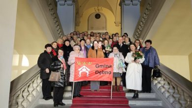 Członkowie klubów seniora pozują do wspólnego zdjęcia na reprezentacyjnych schodach budynku, trzymając czerwony baner z napisem „Gmina Dębno”; część osób trzyma kwiaty i upominki.