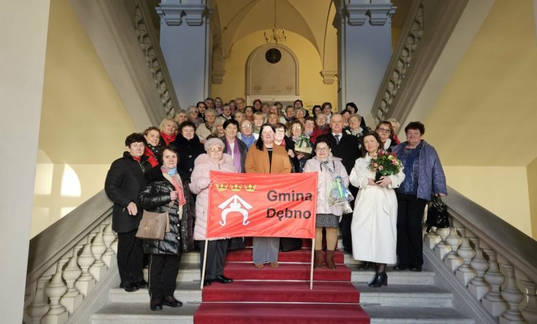 Członkowie klubów seniora pozują do wspólnego zdjęcia na reprezentacyjnych schodach budynku, trzymając czerwony baner z napisem „Gmina Dębno”; część osób trzyma kwiaty i upominki.