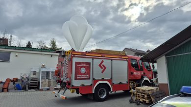 Wóz strażacki zaparkowany na podwórzu przy budynkach gospodarczych, z widocznym sprzętem na dachu i paletami obok. Nad samochodem unosi się duży biały balon meteorologiczny na tle pochmurnego nieba.