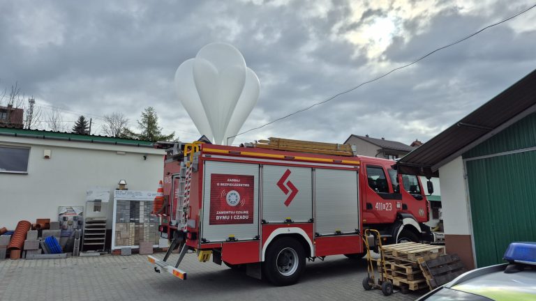 Wóz strażacki zaparkowany na podwórzu przy budynkach gospodarczych, z widocznym sprzętem na dachu i paletami obok. Nad samochodem unosi się duży biały balon meteorologiczny na tle pochmurnego nieba.
