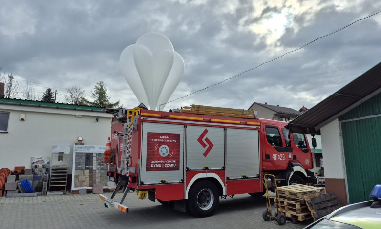 Wóz strażacki zaparkowany na podwórzu przy budynkach gospodarczych, z widocznym sprzętem na dachu i paletami obok. Nad samochodem unosi się duży biały balon meteorologiczny na tle pochmurnego nieba.